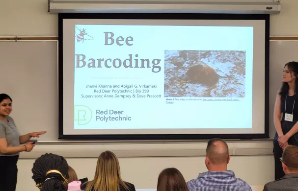 Two female students stand in front of an audience with a presentation being projected called "Bee Barcoding"