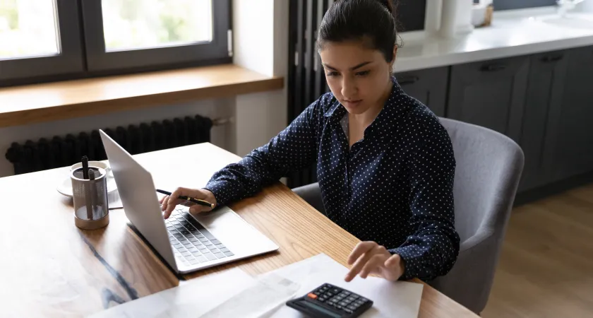 Young woman working on her laptop while entering digits on a calculator.