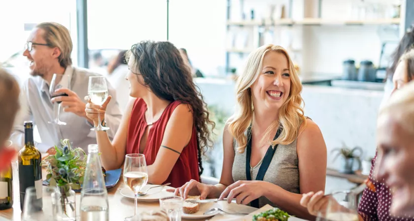 Group of adults sitting around a table smiling, eating food and drinking wine.