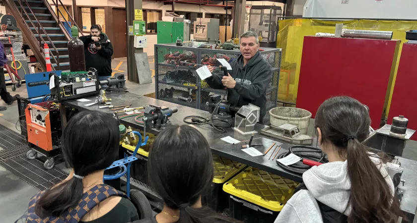 A male instructor stands behind a desk full of tools explaining how they work to a group of three female students