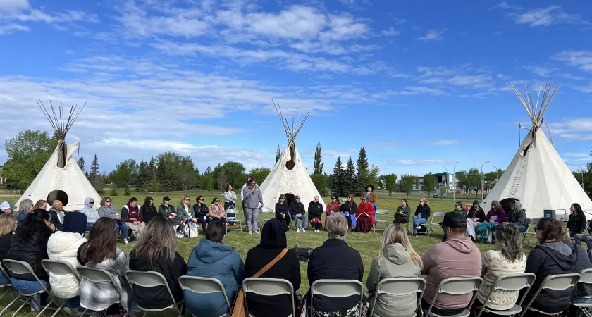 A group sits outside in a large circle on a sunny day. Behind them are three teepees