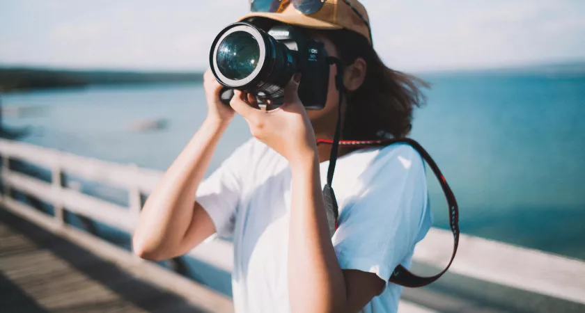Someone in a hat holds a large camera in front of their faces while standing on a pier