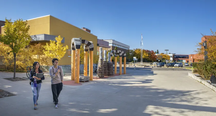 One male and one female student walk outside RDP's library entrance. The leaves on the trees in the background are turning yellow