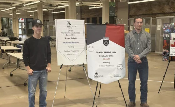 Matthew Fenton stands smiling with his hands in his pockets next to a banner. On the right, Ben Rainforth stands smiling with his hands crossed beside another banner