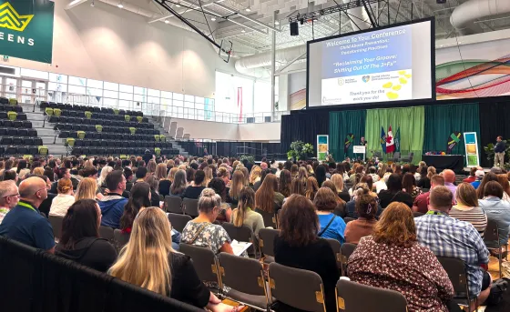 A large crowd sits in the GW Harris main gym looking towards a keynote speaker who's standing on the main stage