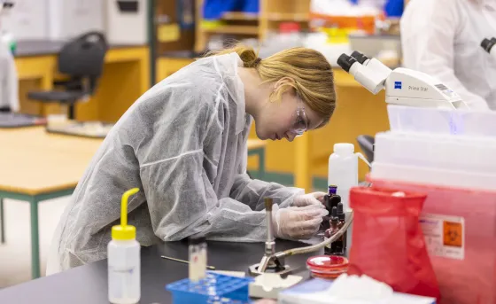 A female student in protective gear works with small bottles of chemical compounds in a lab