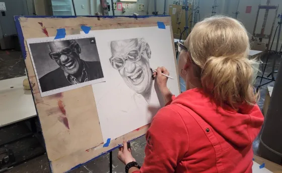 A woman in a red hoodie sits in an art studio. She is sketching a picture of Ray Charles using a pencil