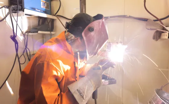Student in orange coveralls and a welding facemask welds in a shop