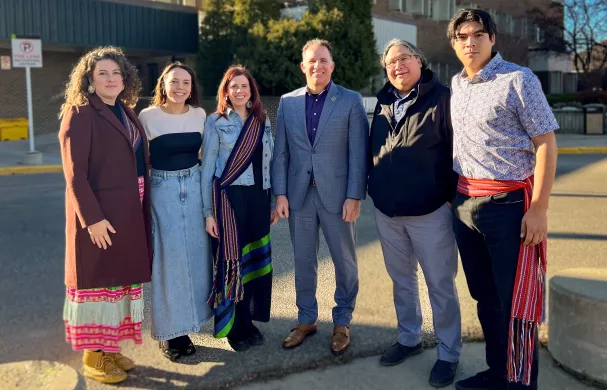 A group of students, admin, and community members stand together. Two wear the Métis Sash and two wear Ribbon Skirts.