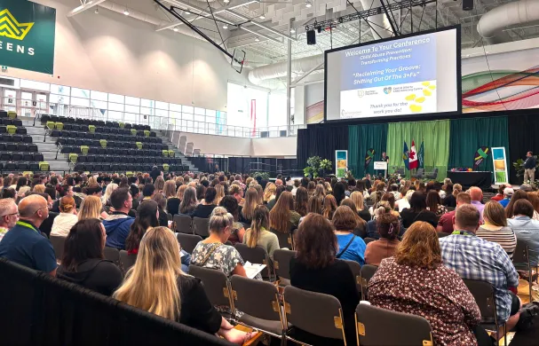 A large crowd sits in the GW Harris main gym looking towards a keynote speaker who's standing on the main stage