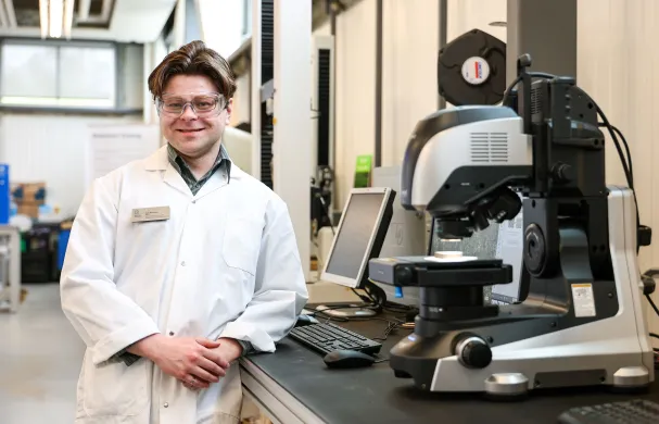 A man in a lab coat and safety glasses leans on a desk that contains a computer and some futuristic looking equipment