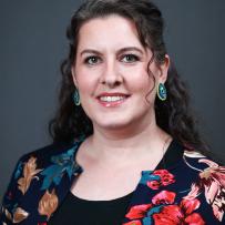 Katherine Thompson has dark wavy hair and wars a floral blouse. She is smiling against a dark backdrop