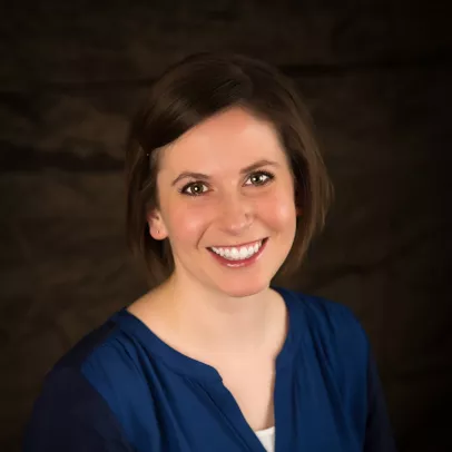 Sabrina smiles at the camera in a professional photo taken against a dark brown backdrop. Her short brown hair is held by a pin on the side. She is wearing a blue shirt with white camisole.