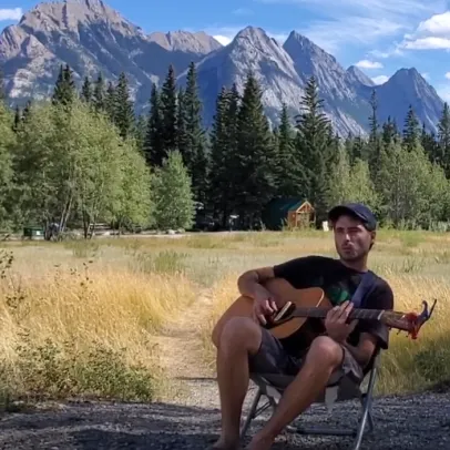 Etienne LeBel sits in a lawn chair playing a guitar. There is a beautiful mountainscape behind him.