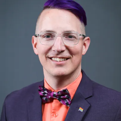 Robbie has purple hair swept to one side, glasses, and wears a purple suit, bright orange shirt, and purple bowtie.