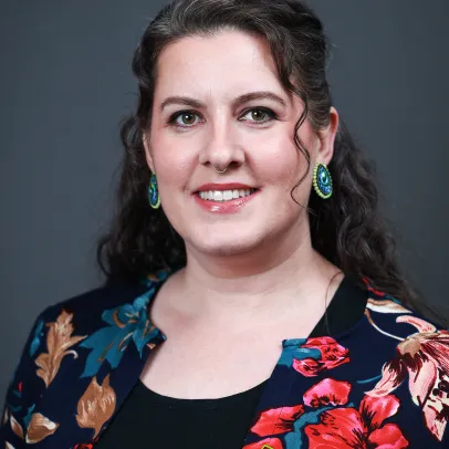 Katherine Thompson has dark wavy hair and wars a floral blouse. She is smiling against a dark backdrop