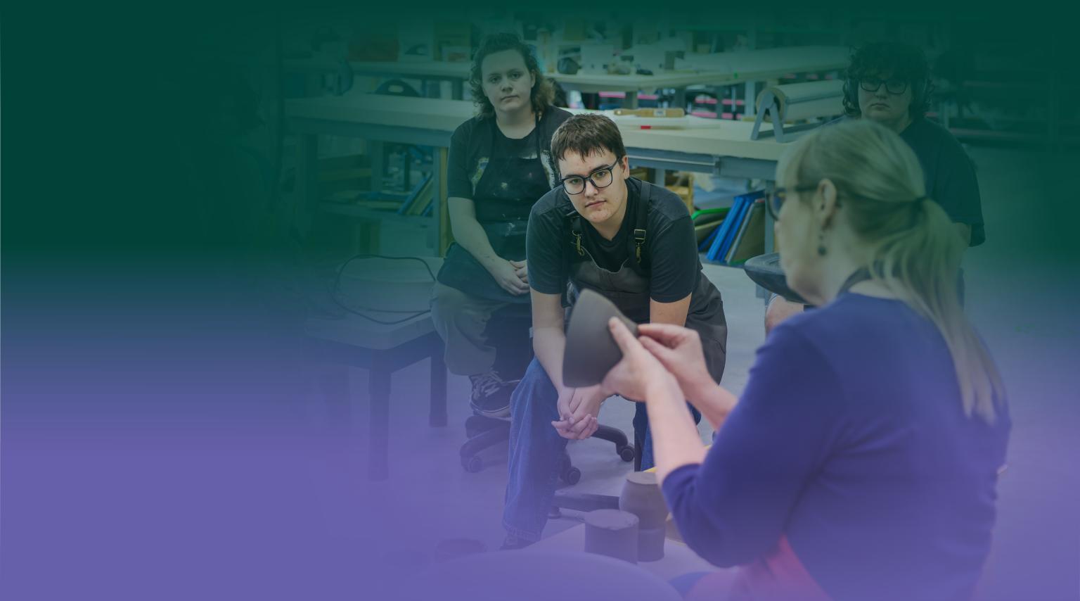 Two students in a pottery studio look towards an instructor who is demonstrating techniques with clay