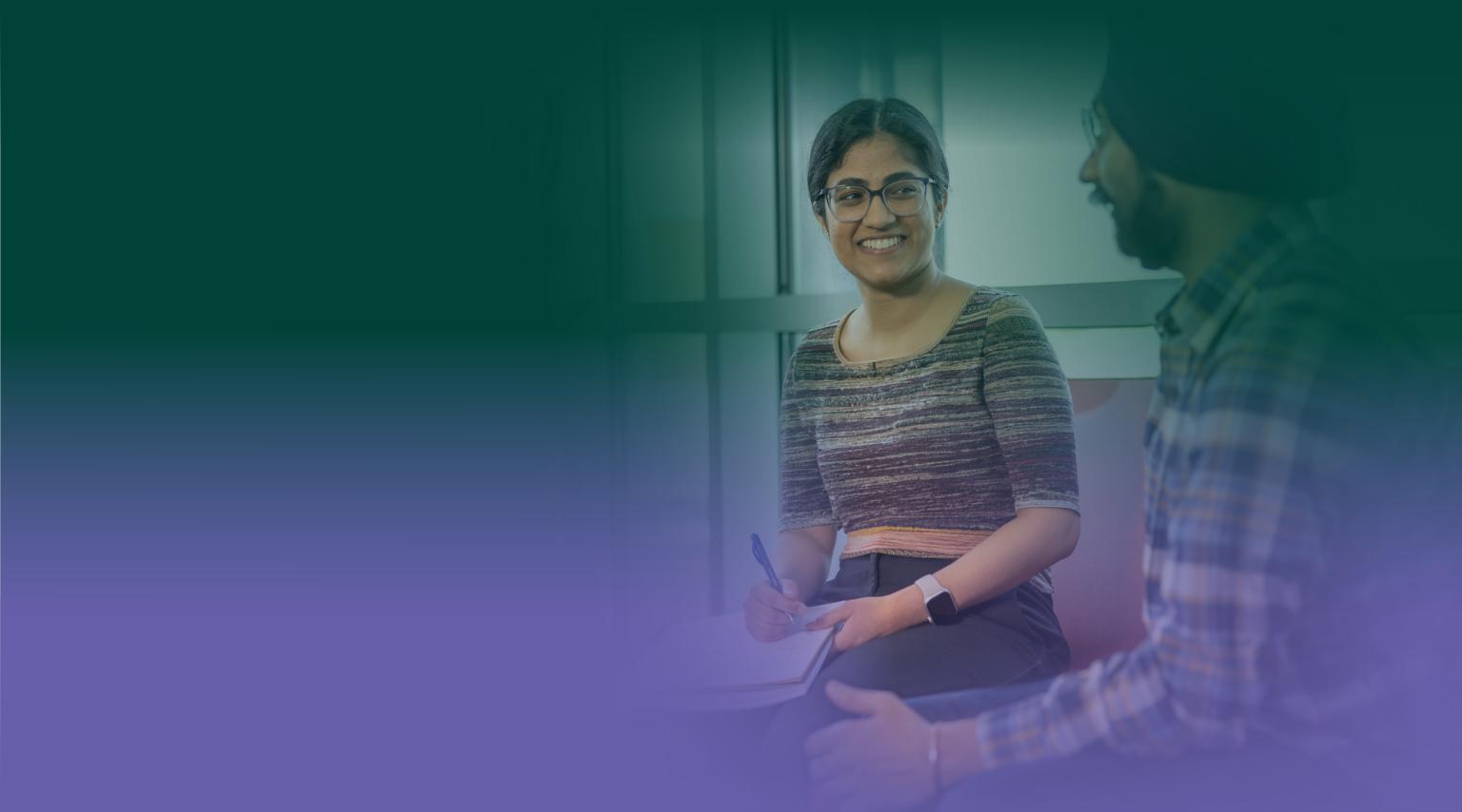 A female student sits in the library with a pen and pad of paper. She's smiling at a Sikh male student who's chatting with her