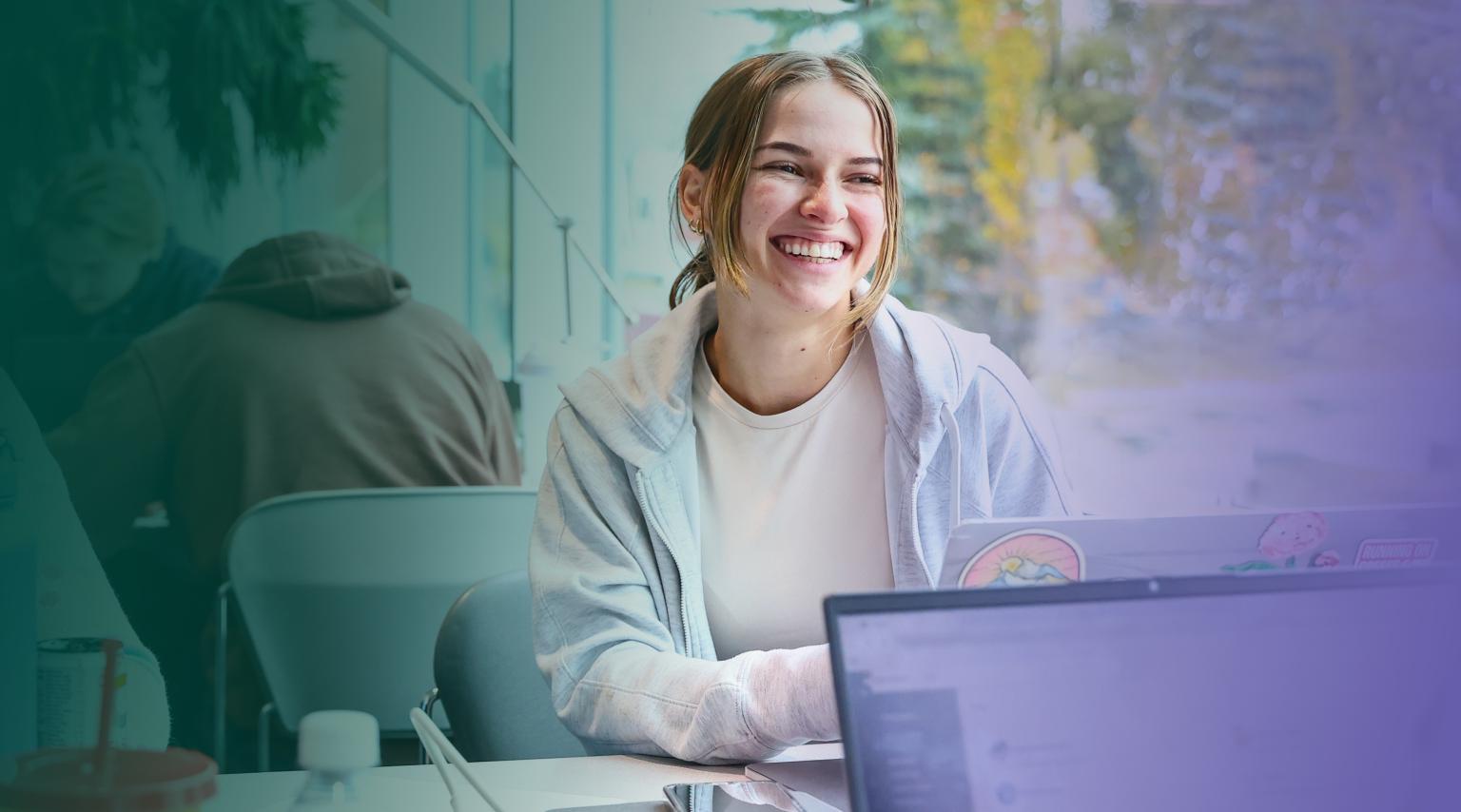 A smiling female student sits at a table with a laptop open in front of bright windows