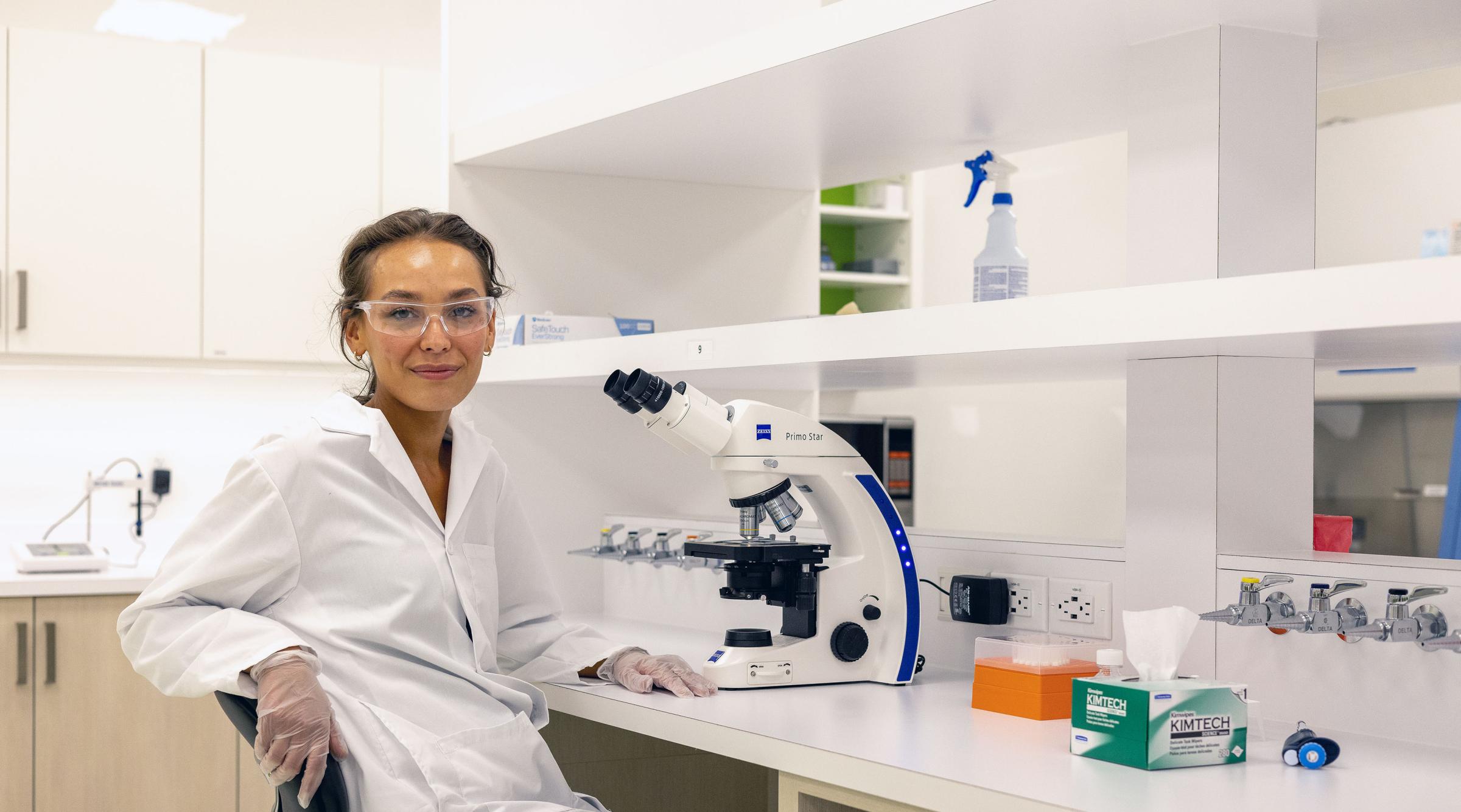 A researcher turns towards the camera, leaning their arm on the back of their chair, beside them is a microscope and various bottles.