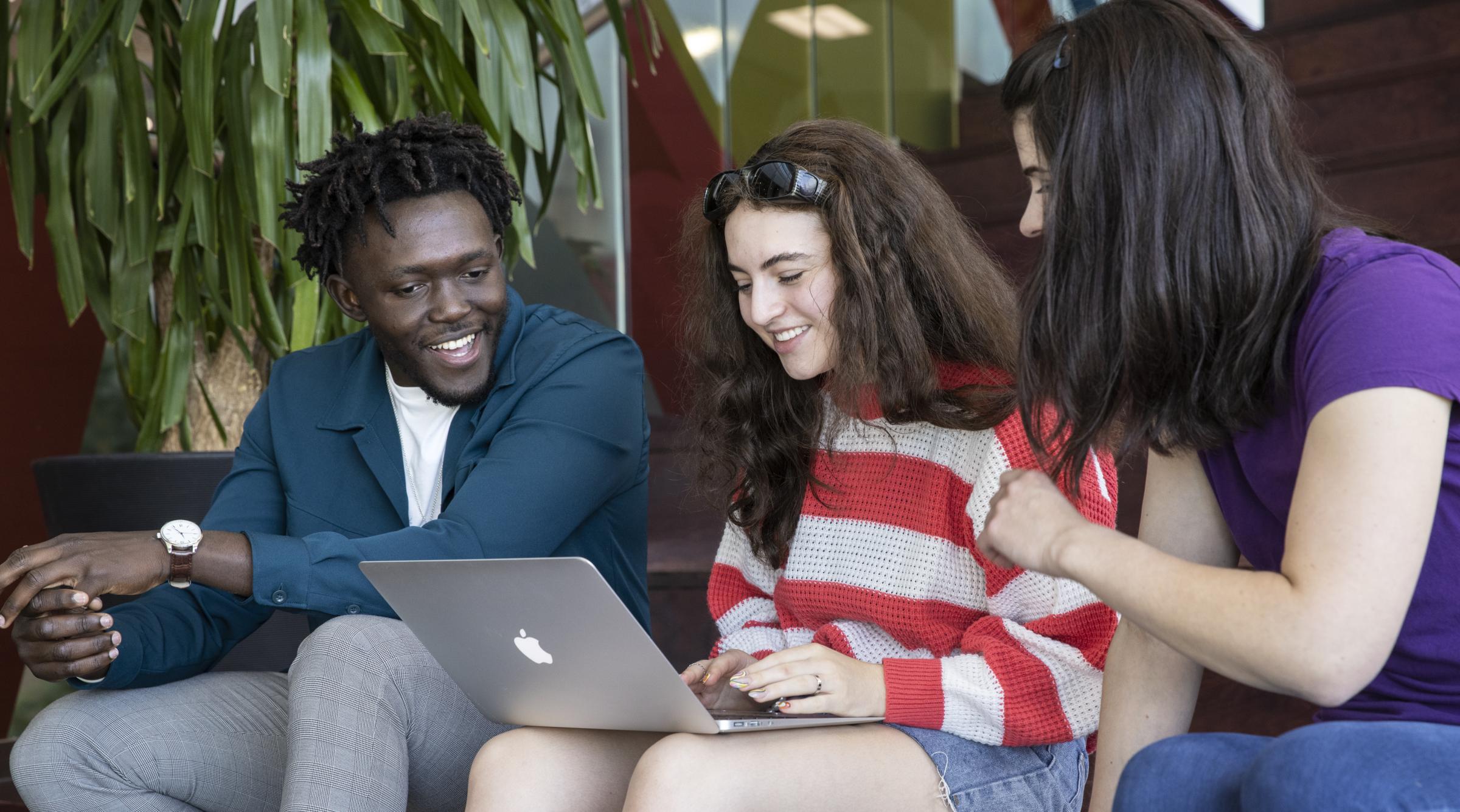 Three smiling students sit on stairs and look at laptop