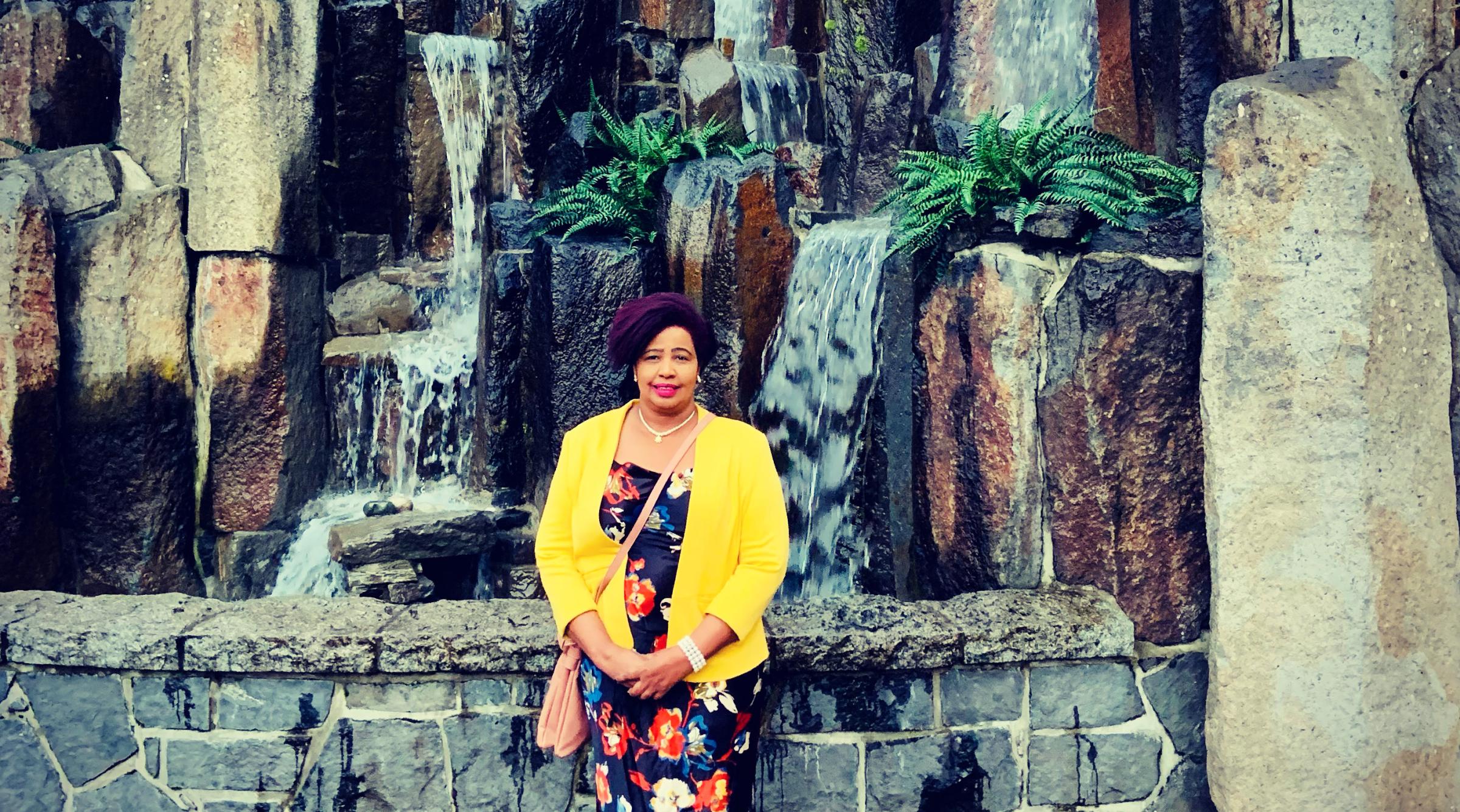 Zanaib stands in front of a large rock formation, wearing a flowered dress and yellow jacket.