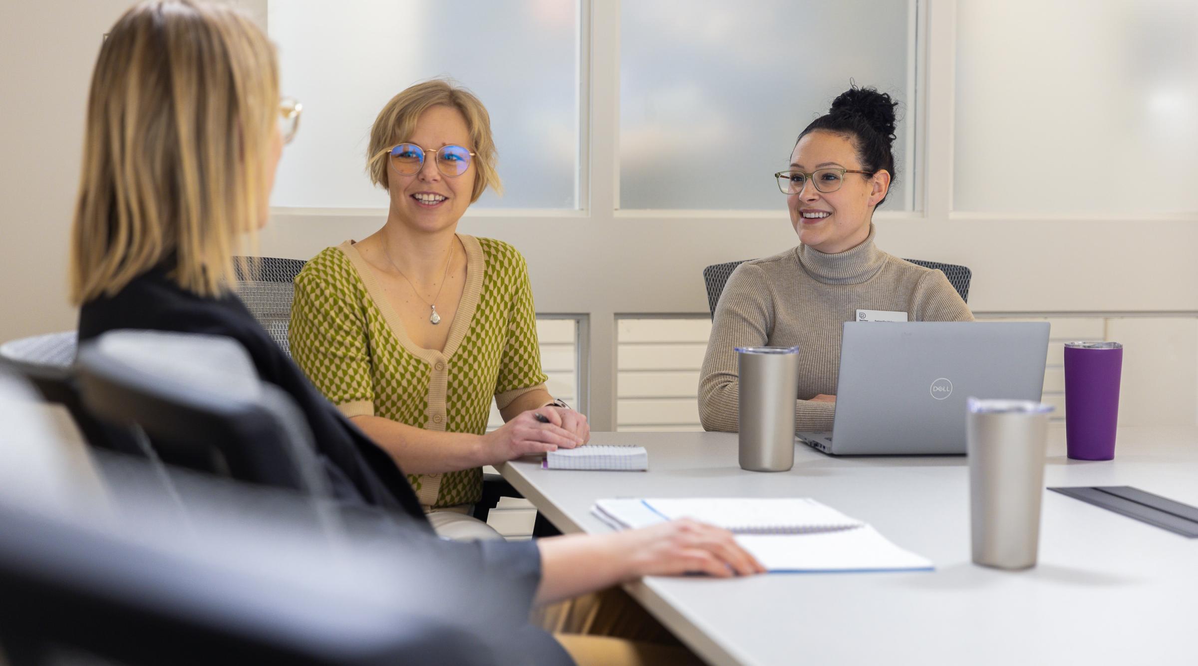 Two women sitting at a table with laptop and notebook looking at a blonde person whose back is to the camera. They appear to be in a meeting.