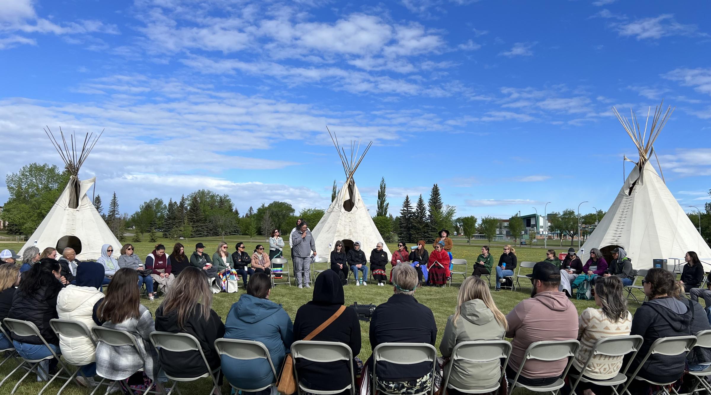 A group sits outside in a large circle on a sunny day. Behind them are three teepees