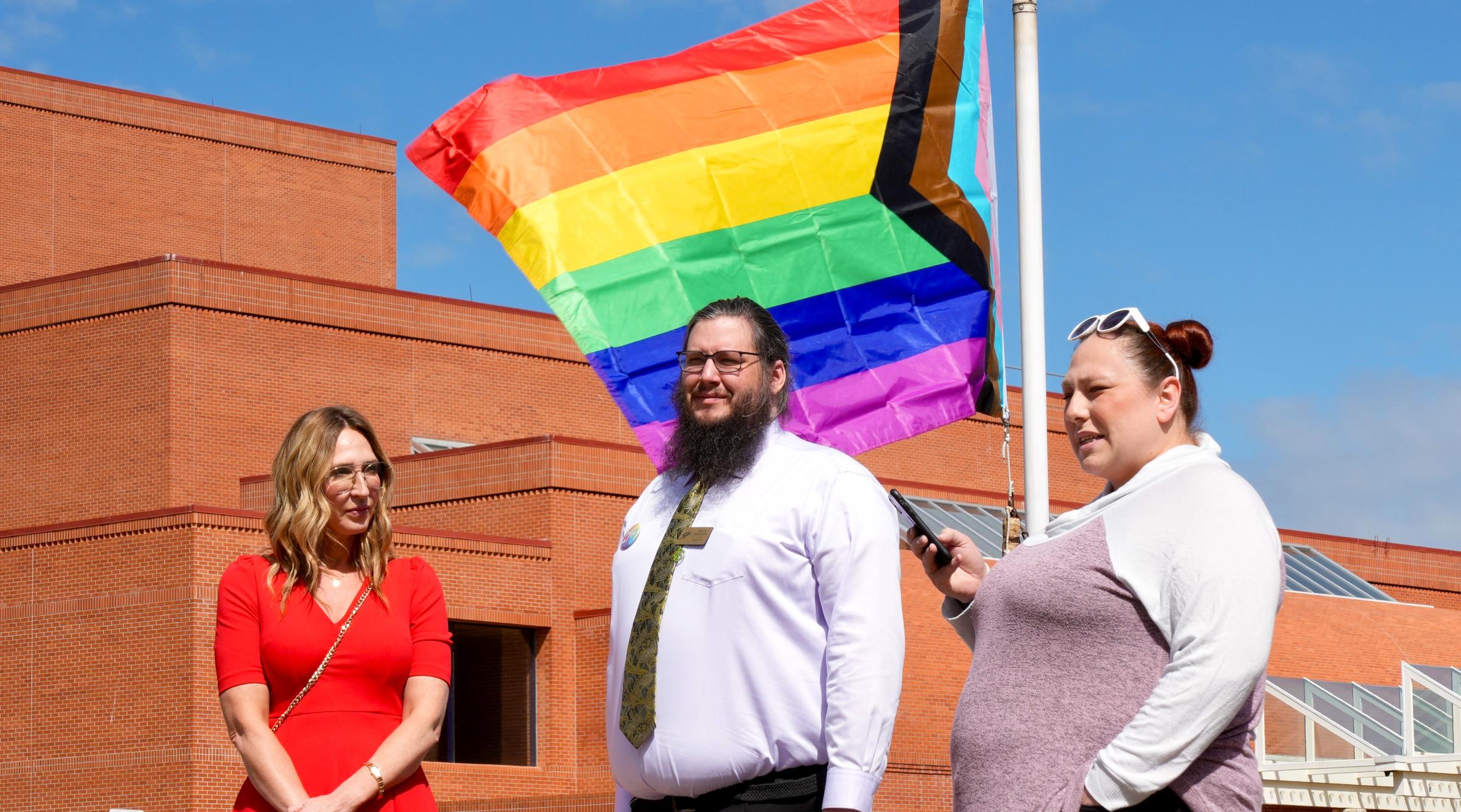 A group of three people stand in front of the Pride flag as it is raised up a flagpole at RDP
