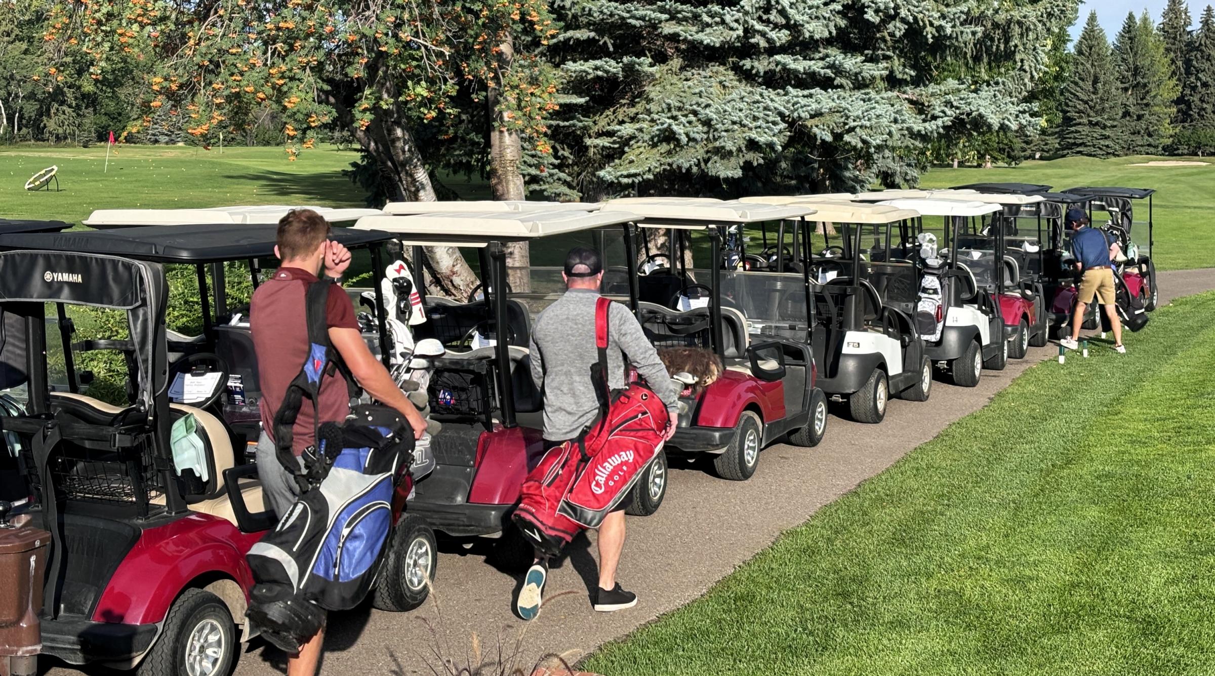 A row of golf carts is lined up next to a fairway. A few golfers walk towards the carts with golf bags on their shoulders