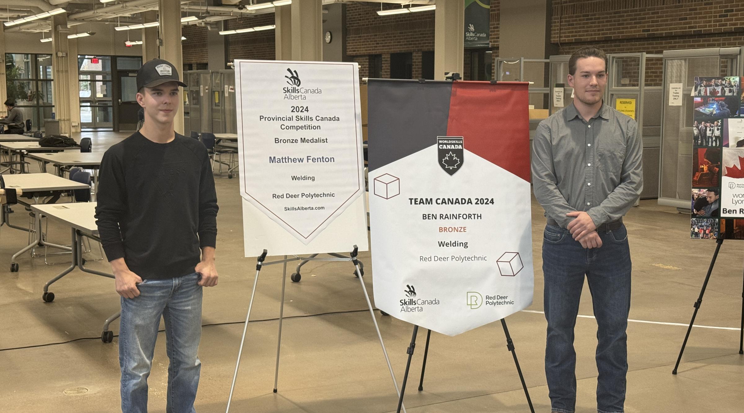 Matthew Fenton stands smiling with his hands in his pockets next to a banner. On the right, Ben Rainforth stands smiling with his hands crossed beside another banner