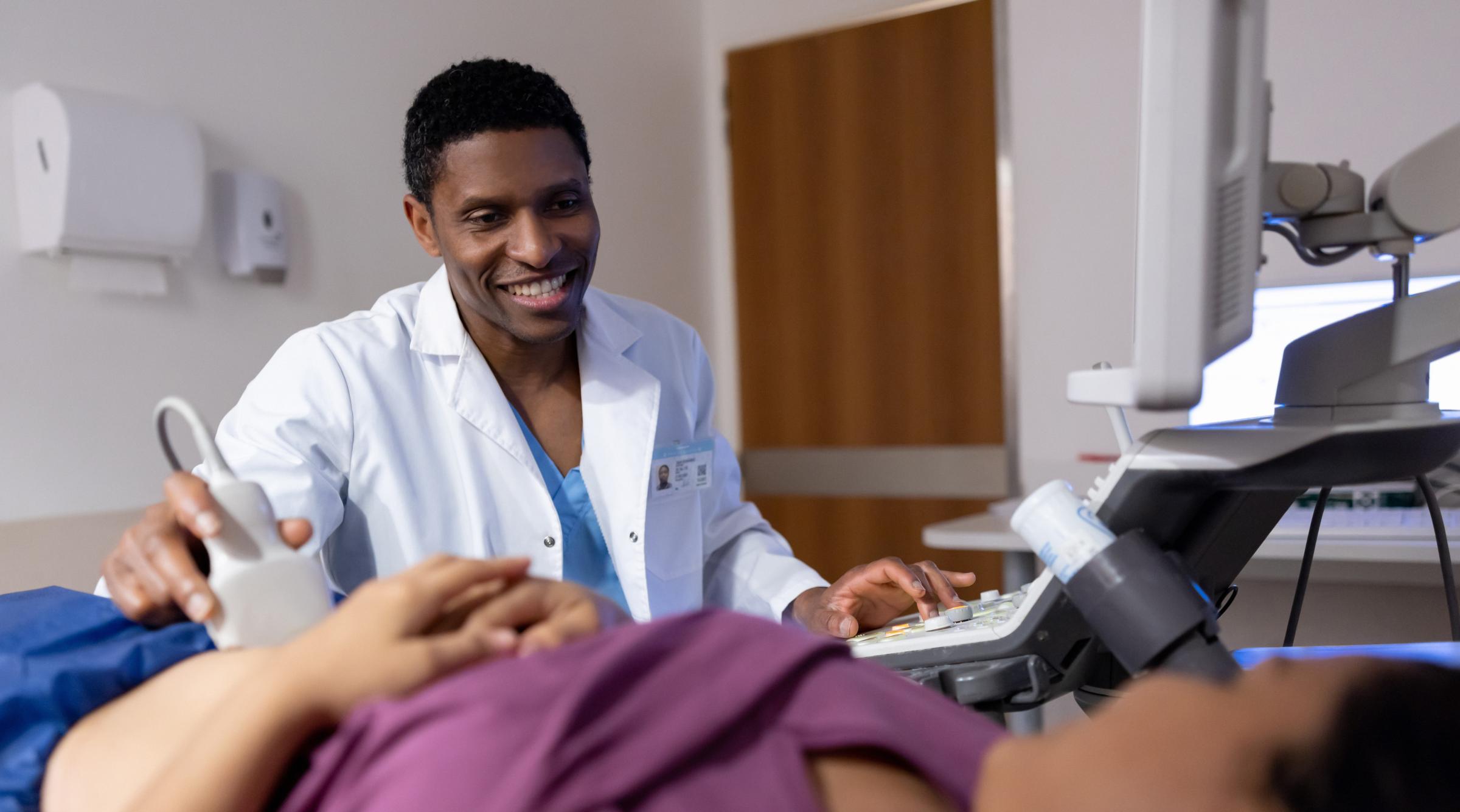 A sonographer smiles while using an ultrasound wand on a patient abdomen.