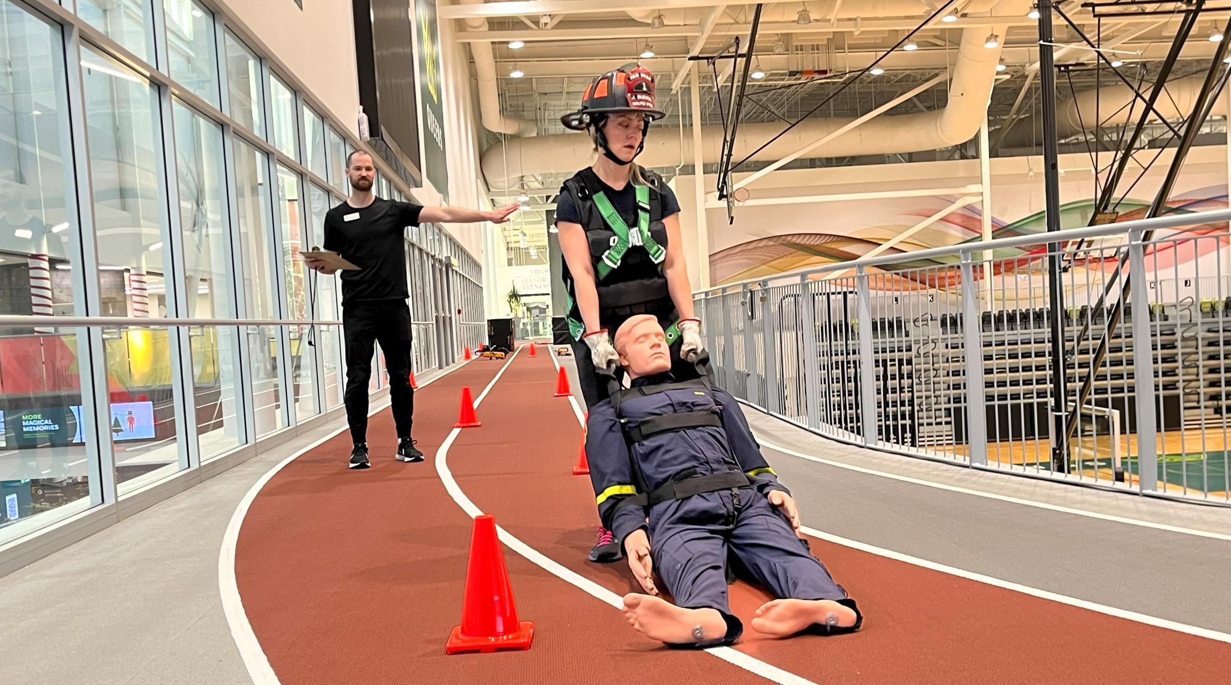 A woman in weighted vest carries a weighted manakin through a course as a personal trainer directs her
