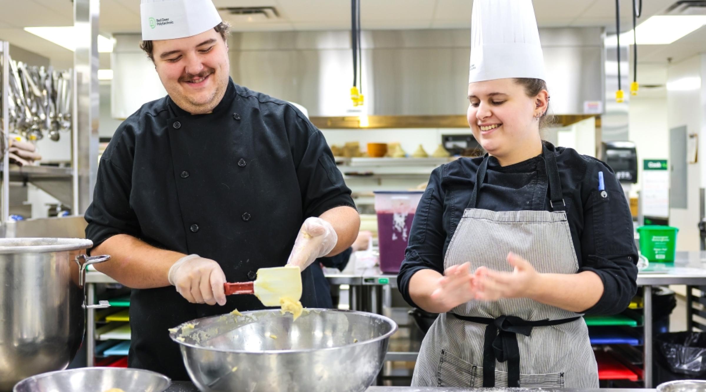 A smiling male and female student in chef's hats prepare some baking in a commercial kitchen