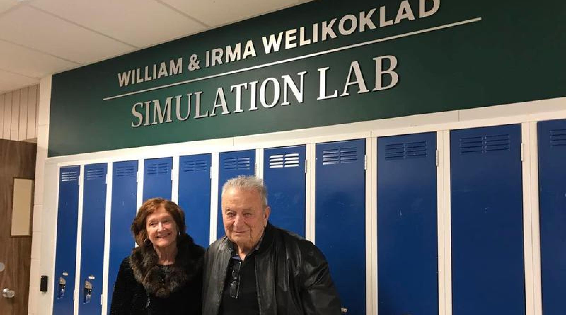 Irma and Bill Welikoklad stand smiling in front of the Welikoklad Simulation Lab