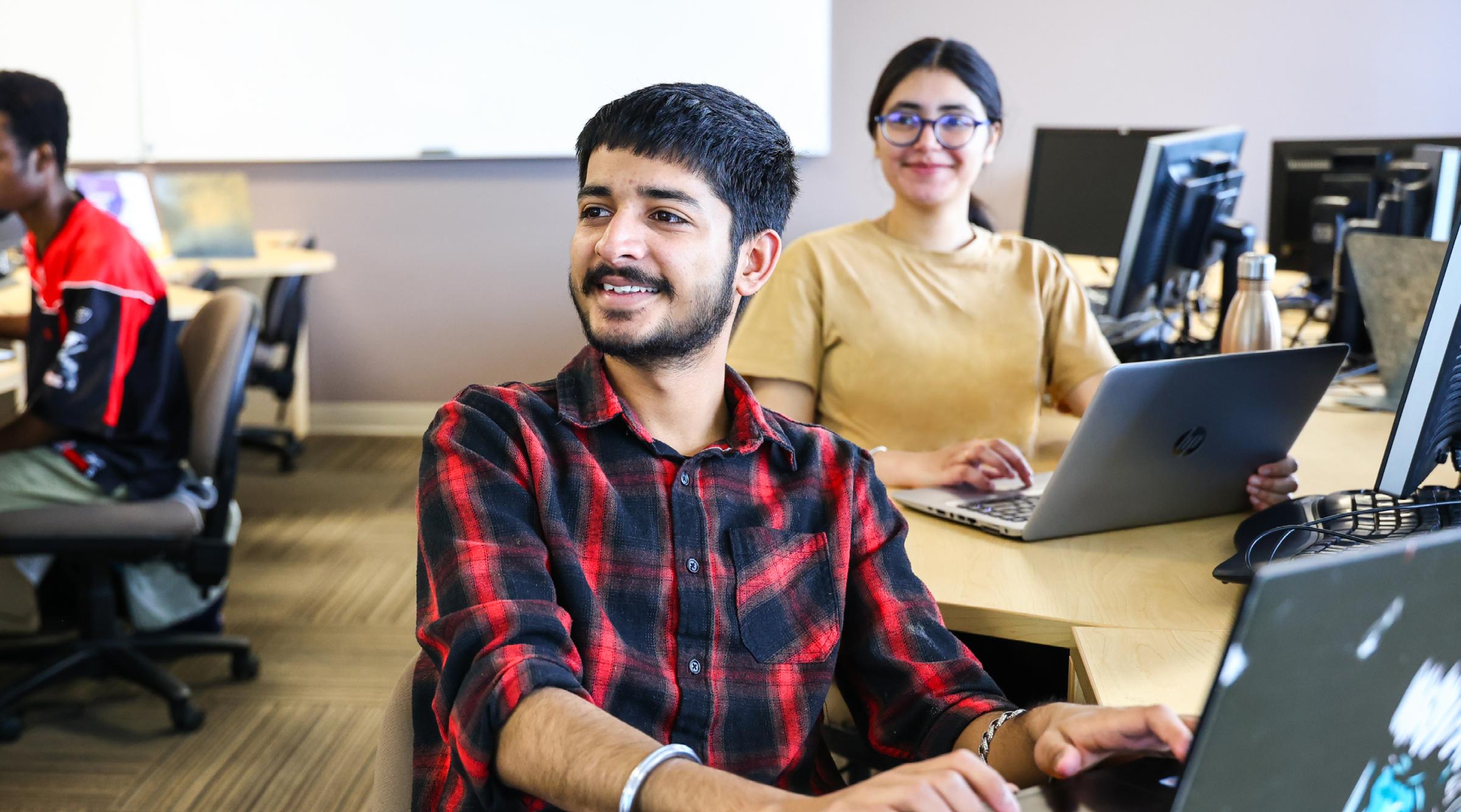 A, student in a plaid shirt, smiles to the instructor (who is not seen) in a computer lab classroom.