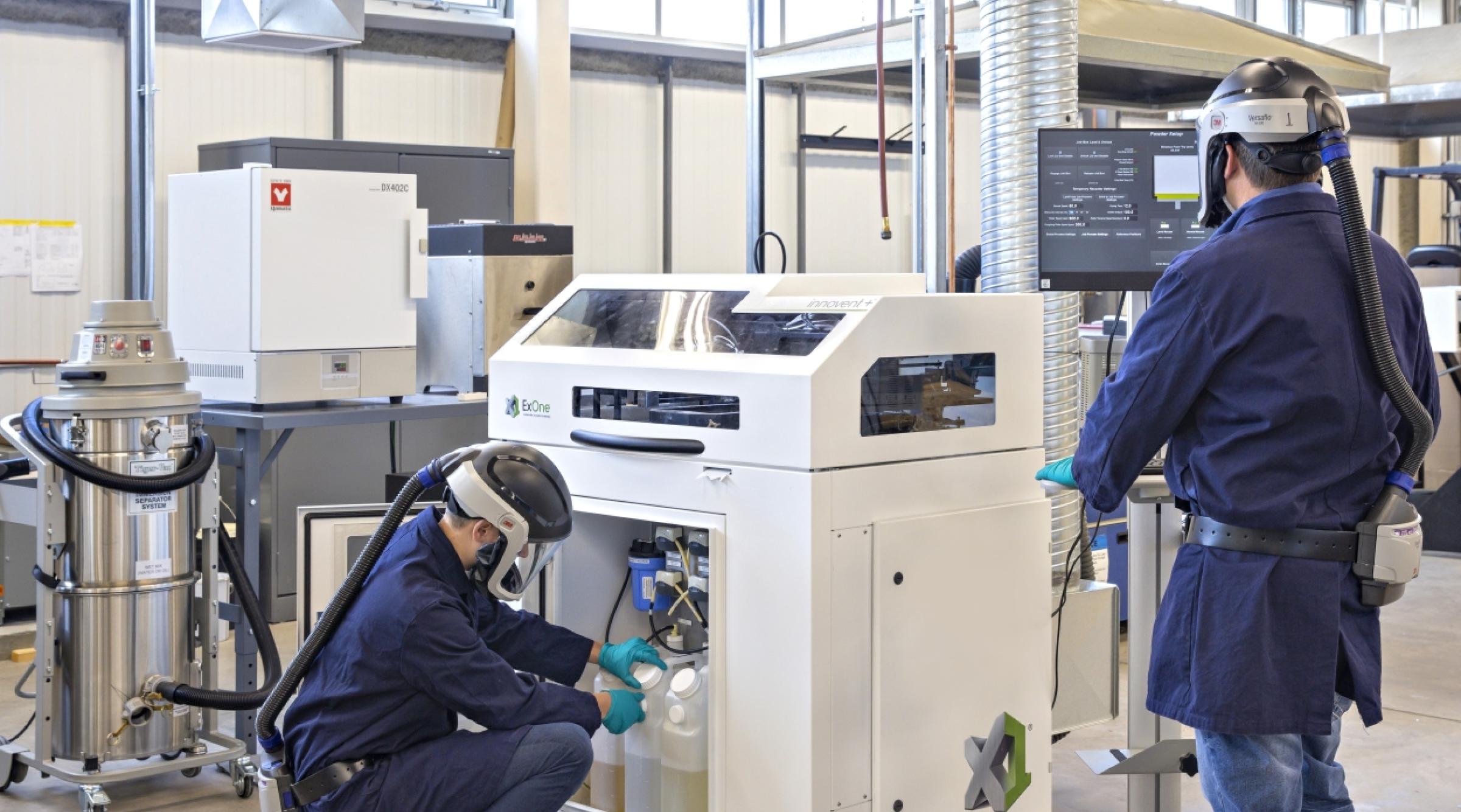 Two men wearing blue lab coats and protective helmets are working with the ExOne 3D Printer
