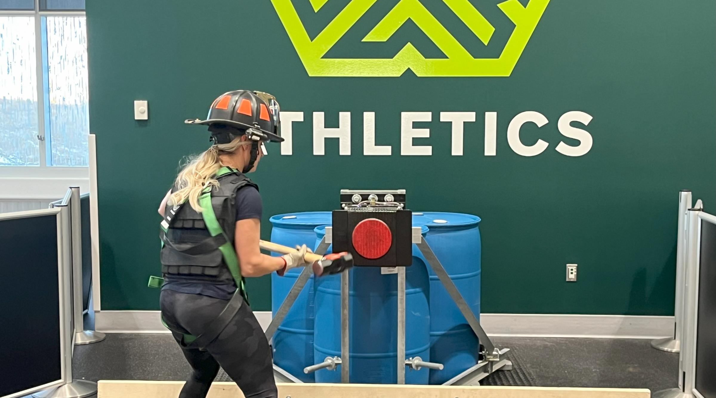 A female in a firefighter helmet and harness is swinging a hammer against a large red target as part of the Firefighter Applicant Physical Test
