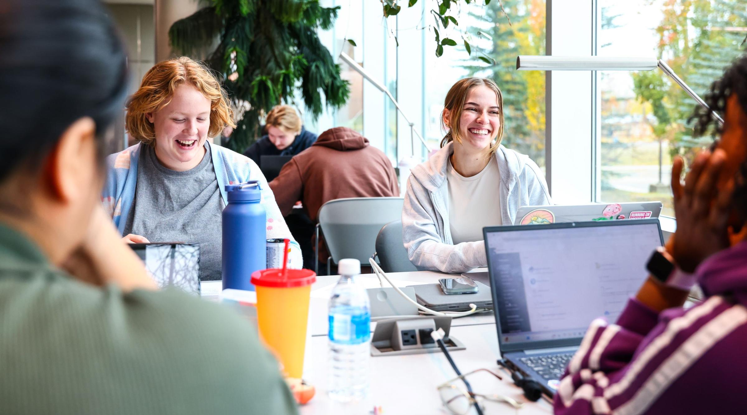 Two smiling female students sit across from a few more students who have their back to the camera