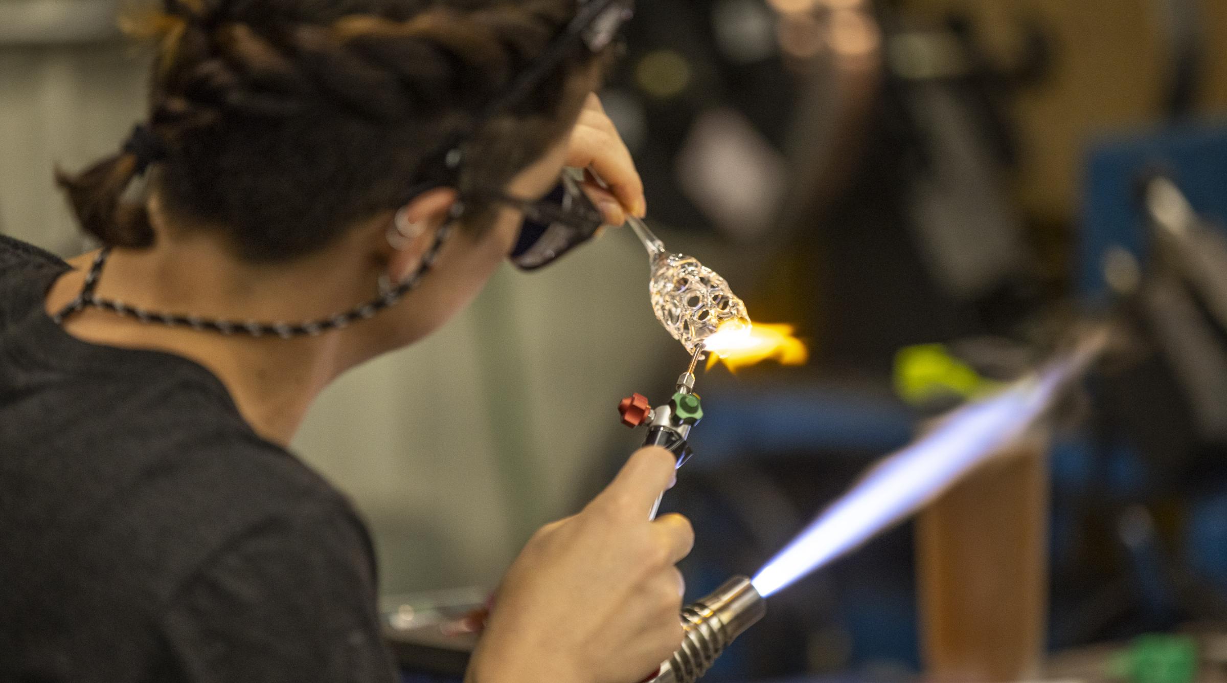 An artist with braided hair uses a torch to create a design in glass.