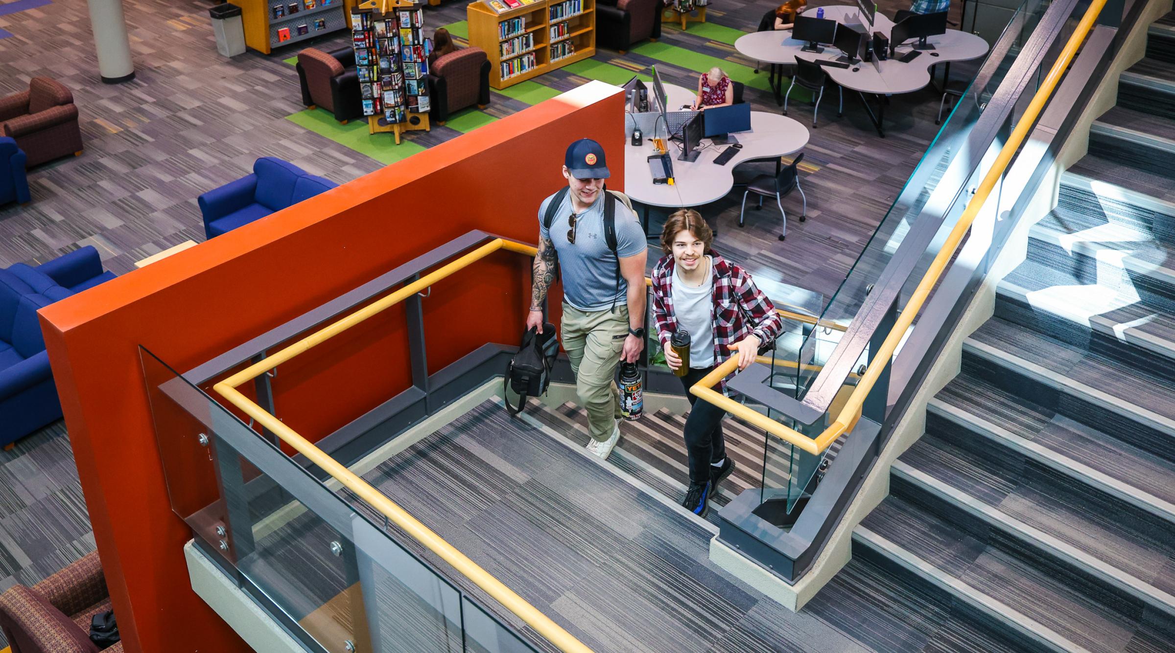 Two students walk up the stairs in the library. Below are shelves of books and students studying at tables with computer screens.