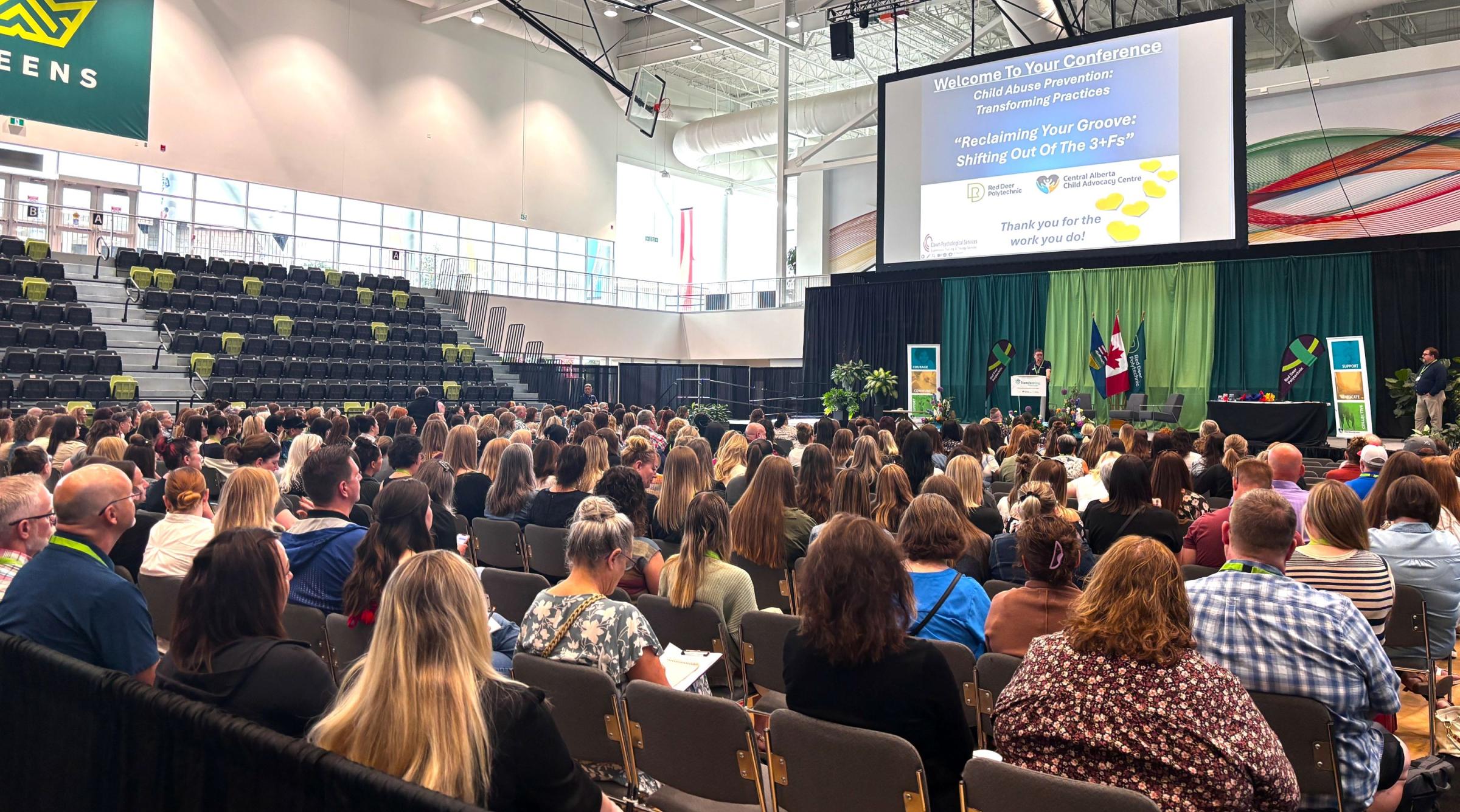 A large crowd sits in the GW Harris main gym looking towards a keynote speaker who's standing on the main stage