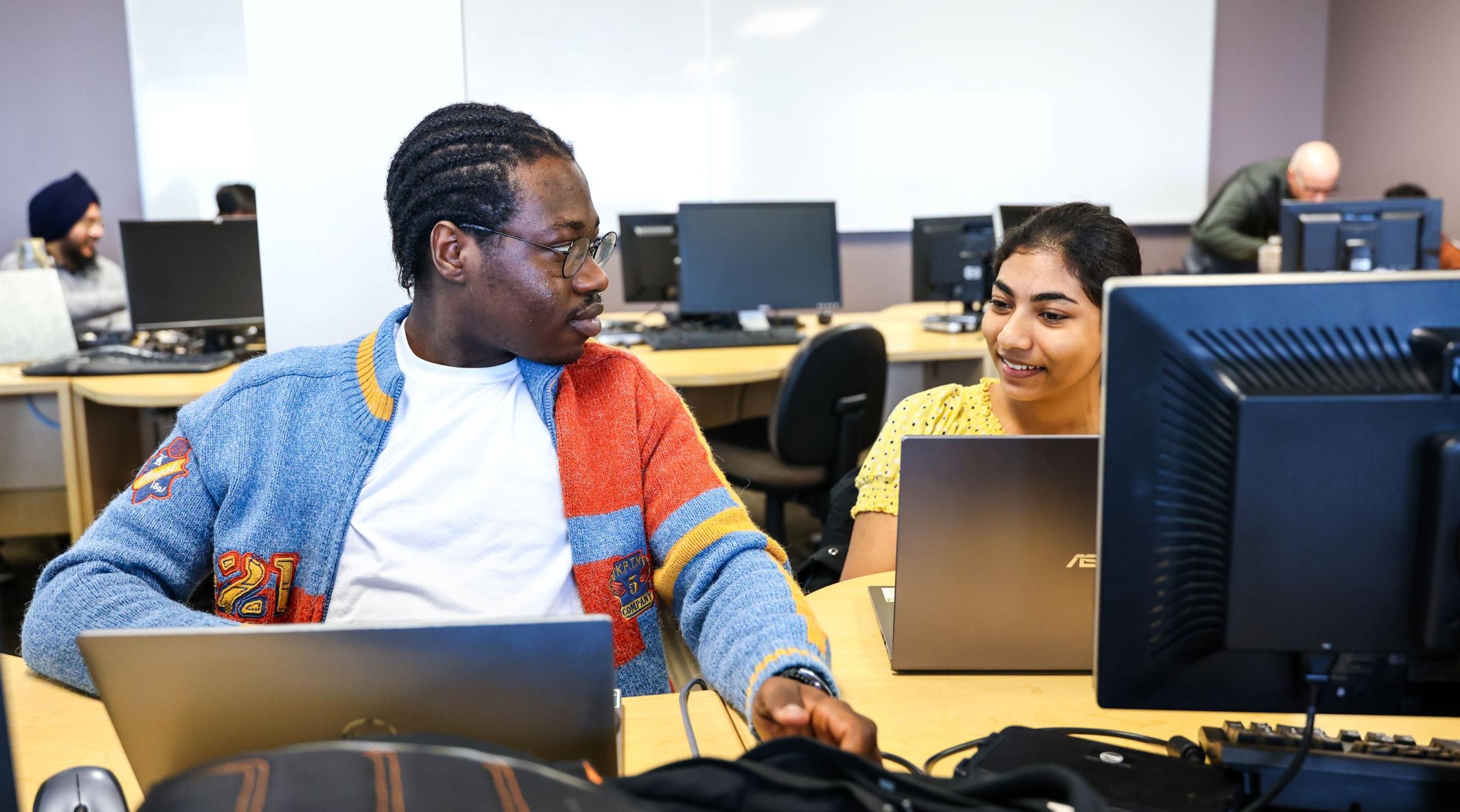 A male and female student sit next to each other in a computer lab. They have laptops in front of them and are smiling at each other