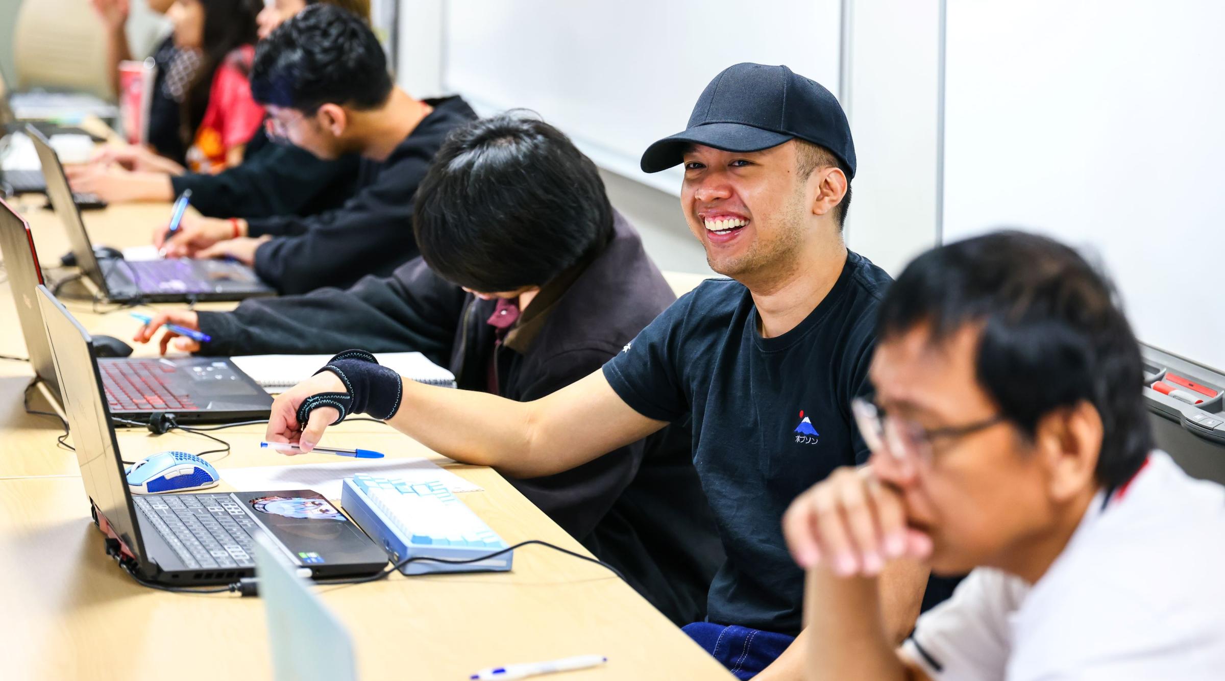 A group of students sit in class in front of their laptops. One young man looks up and smiles