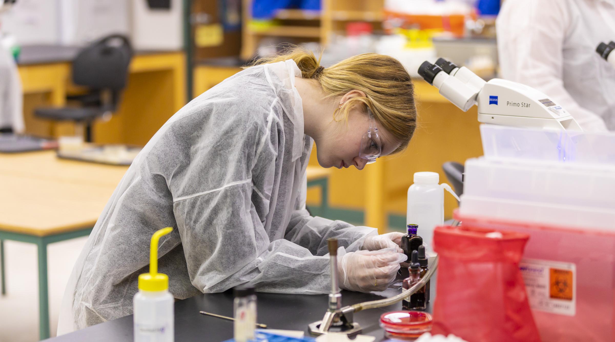 A female student in protective gear works with small bottles of chemical compounds in a lab