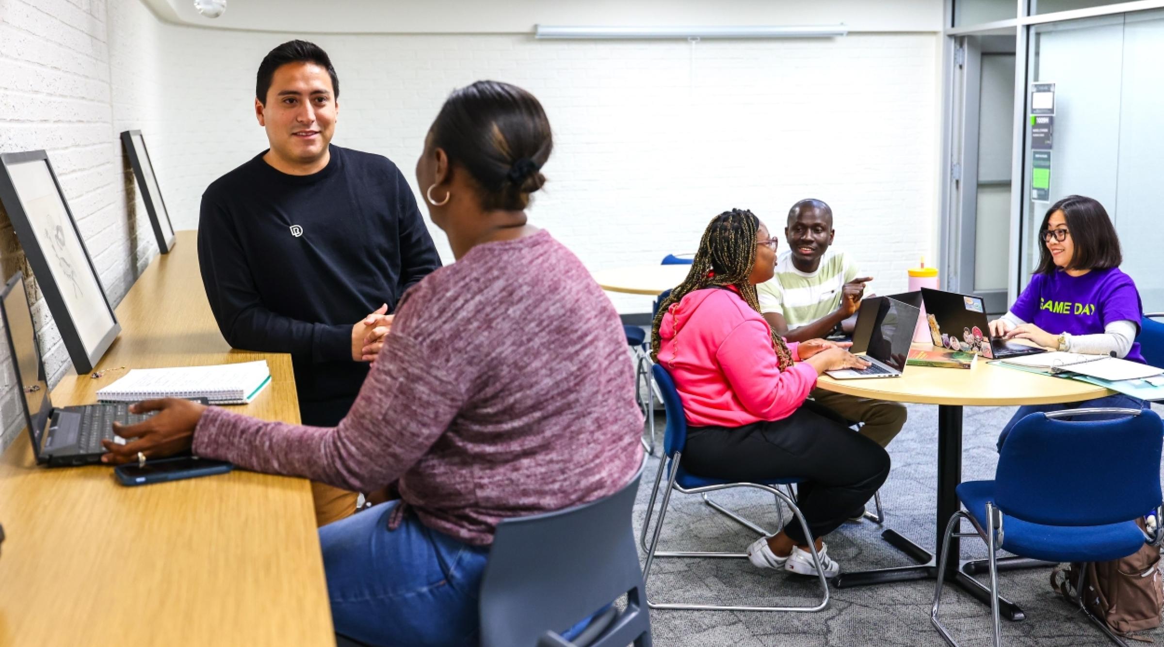 A man chats with a Black woman who has her back turned to the camera. In the background, a group of Black students chats at a round table