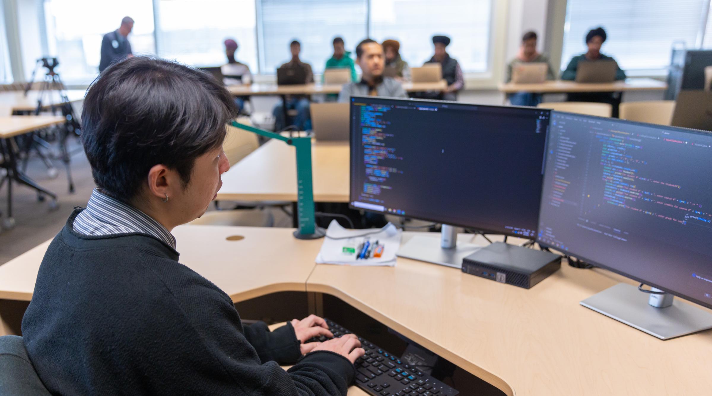 A male student sits coding in front of two screens in a classroom full of fellow students
