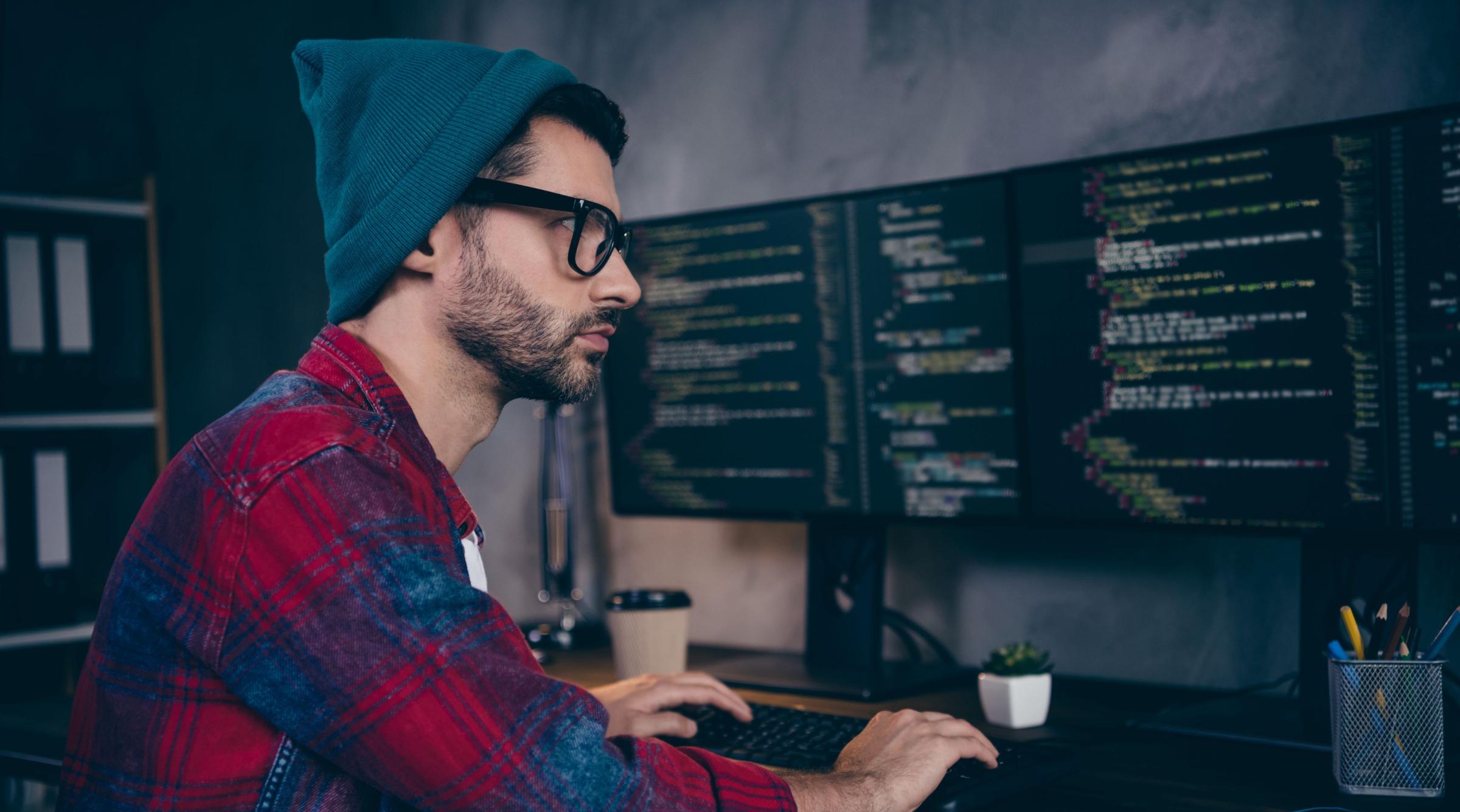 A man in a blue toque, red flannel, with thick glasses sits in front of two computer monitors