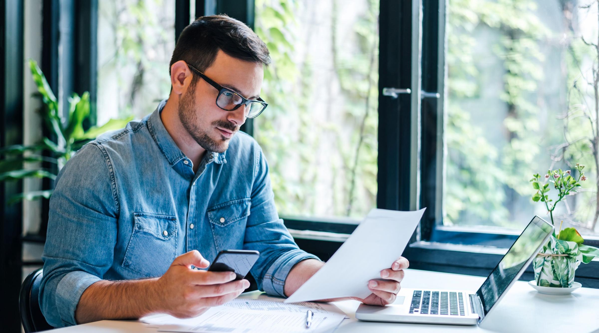 Early professional man in glasses and a denim shirt sits at a desk with his laptop, some papers, and his phone in hand. 