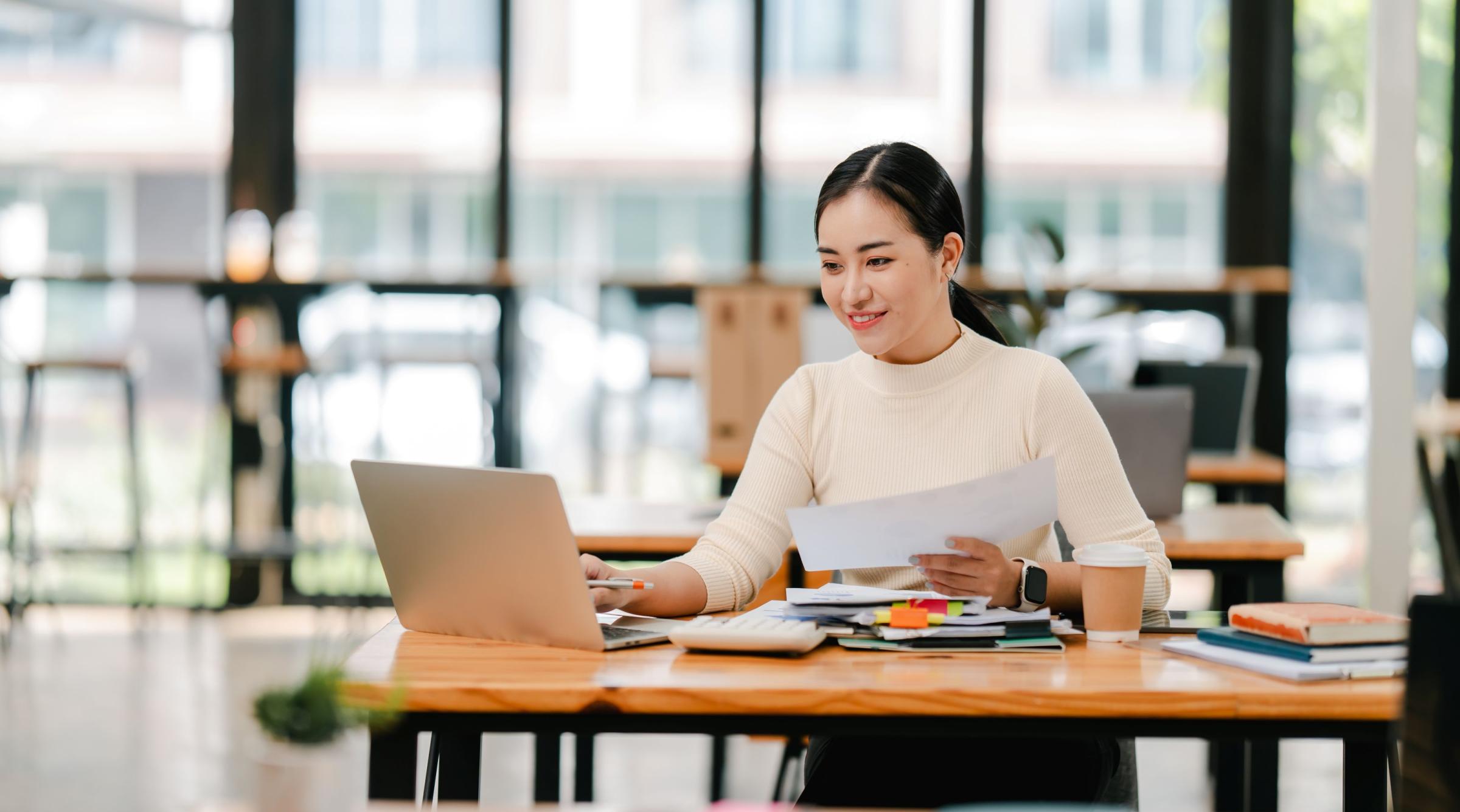 A woman sits in a bright office in front of a table with a laptop and a number of books
